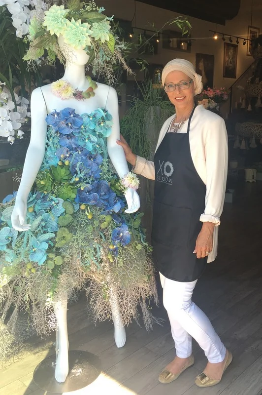 Florist standing beside a mannequin dressed in an artistic floral gown made of blue and green flowers, displayed inside a flower shop as a creative botanical fashion installation.