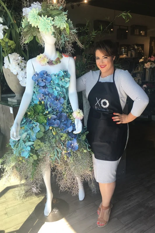 Florist standing beside a mannequin dressed in an artistic floral gown made from blue and green flowers and foliage, displayed inside a flower shop to showcase creative floral design and couture-style arrangements.