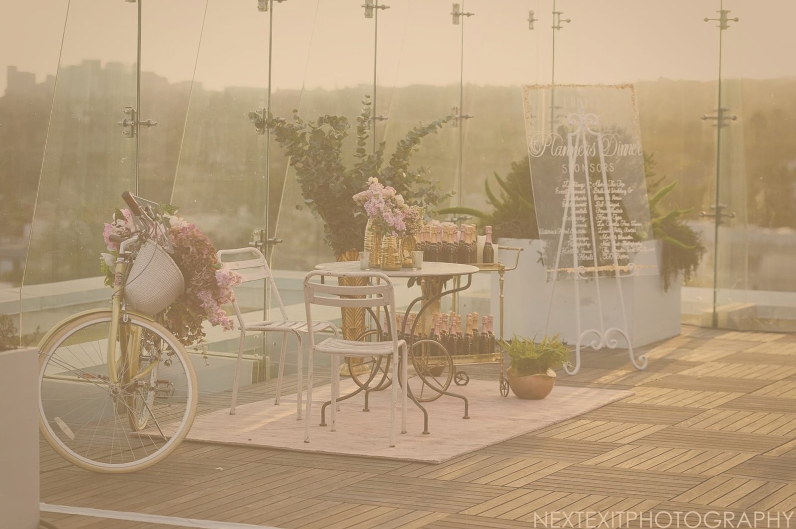 Outdoor event setup featuring a vintage yellow bicycle adorned with flowers, a small table with sparkling beverages, and a decorative sign displaying sponsors, all set against a city skyline backdrop.