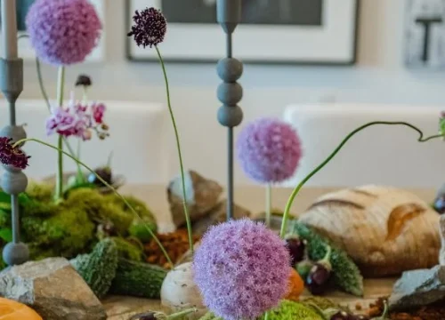 Floral centerpiece featuring vibrant purple alliums and decorative elements, surrounded by greenery and rustic stones, enhancing a luxurious celebration atmosphere.