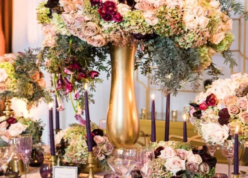Luxurious floral arrangements featuring roses, hydrangeas, and greenery in golden vases, enhancing a celebratory table setting for special events by XO Bloom.