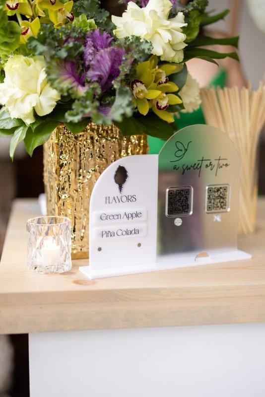 Elegant event tabletop display featuring a floral arrangement, gold vase, candle, and a custom flavour menu sign with QR codes for a luxury cocktail or tasting experience.