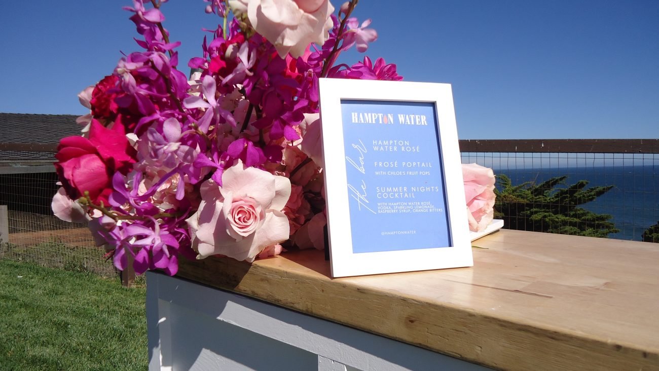 Floral arrangement with pink and purple flowers beside a framed Hampton Water cocktail menu on an outdoor bar, overlooking the ocean at a sunny coastal summer event.