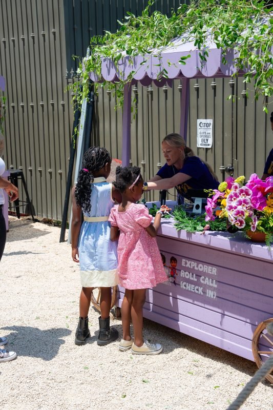 Two young girls stand at a pastel purple outdoor check-in booth decorated with green vines and bright flowers, while a staff member assists them during a family-friendly community event on a sunny day