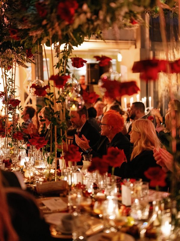 Luxury evening banquet featuring long communal tables, red floral installations, hanging flowers, and candlelight with guests dining in an elegant indoor venue.