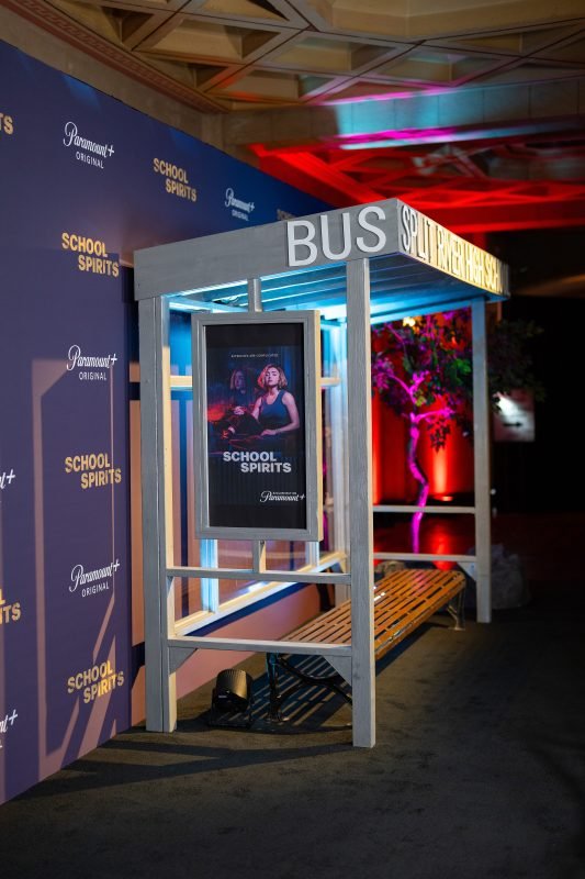 Branded bus stop&ndash;style photo installation for the Paramount+ series School Spirits, featuring illuminated signage, bench seating, and promotional poster at a red carpet or media launch event.
