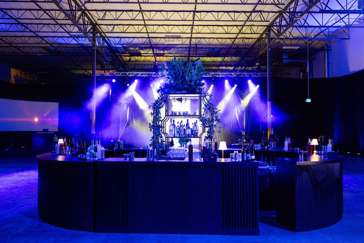 Luxury indoor event bar setup with dramatic blue and purple stage lighting, featuring a central illuminated back bar with bottles and greenery, designed for a high-end corporate or nightlife event.